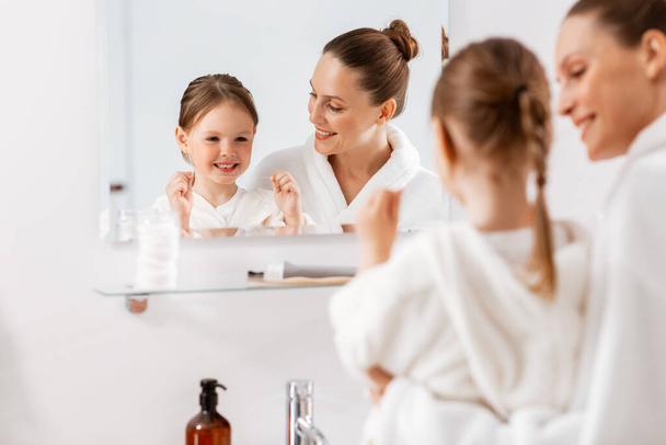 beauty, hygiene, morning and people concept - happy smiling mother and little daughter with dental floss cleaning teeth and looking to mirror at bathroom - Foto, imagen