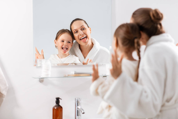 beauty, family and people concept - happy smiling mother and little daughter looking to mirror and hugging in bathroom - Φωτογραφία, εικόνα