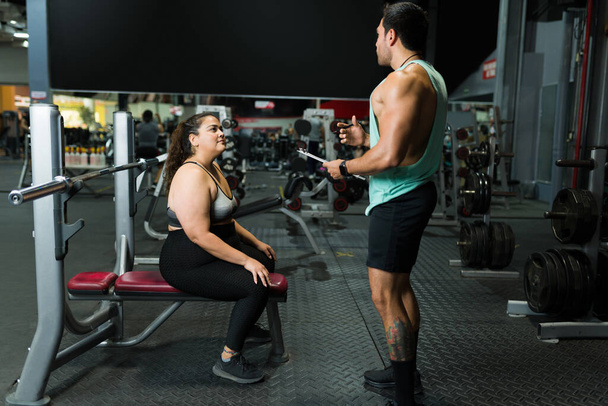 Plus la taille de la femme à la salle de gym en écoutant son entraîneur de conditionnement physique pour commencer une routine d'entraînement pour perdre du poids  - Photo, image
