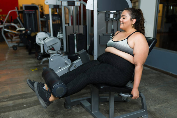 Neşeli şişman kadın spor makinesiyle bacak egzersizi yapıyor. Şişman kadın kilo verme sürecinden memnun. - Fotoğraf, Görsel