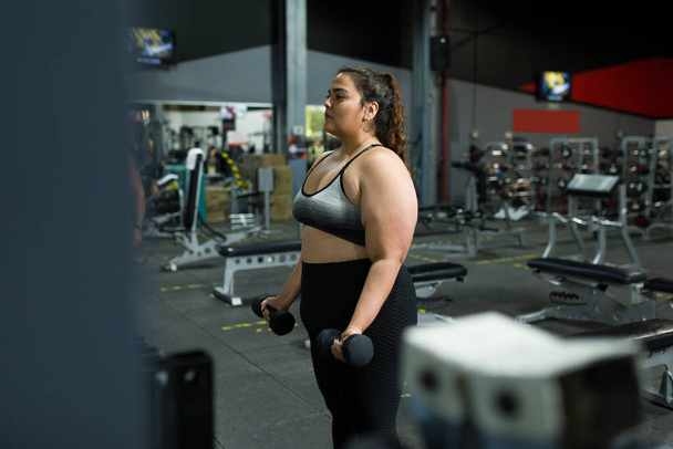 Päättynyt ylipainoinen nainen katselee peiliin kuntosalilla tehdessään painonnosto  - Valokuva, kuva