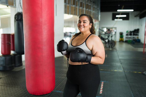 Alegre mujer de talla grande sonriendo y mirando a la cámara mientras practica box en el gimnasio - Foto, Imagen