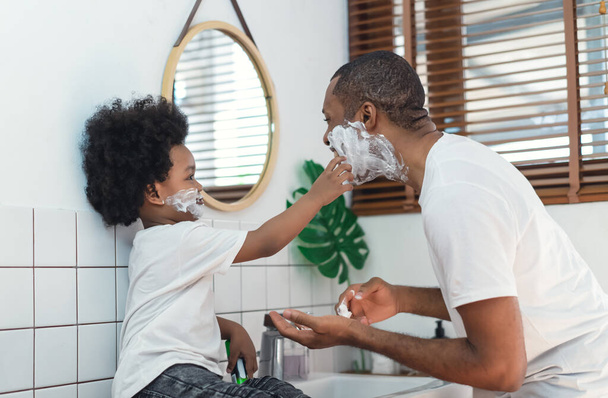 Happy African black father and little son with shaving foam on their faces looking at each other and laughing in the bathroom. Black family spend time together. Morning routine in bathroom concept. - Foto, imagen