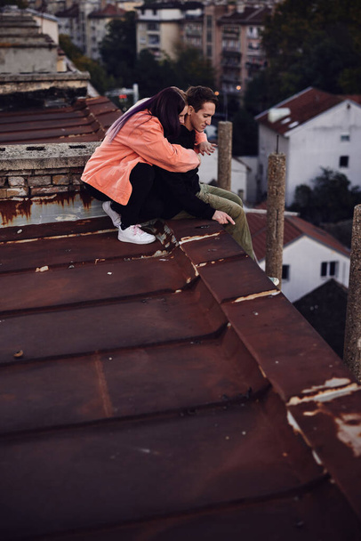 A teenage couple is sitting on top of the building, looking at the view and hugging. - Foto, afbeelding