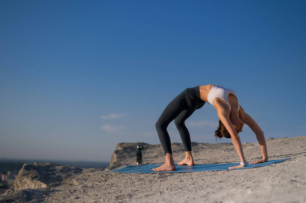Beautiful slender Caucasian girl practices yoga on a rock against the sky and performs the gymnastic bridge exercise - Photo, Image
