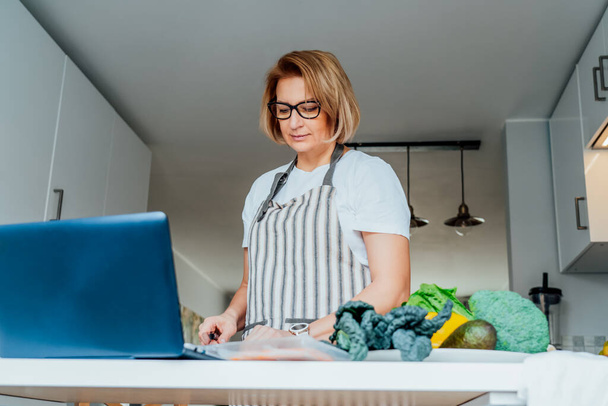 Middle age woman following a cooking tutorial video course on laptop while preparing meal in a kitchen. Woman cooking healthy dish, fish and vegetables on the table. Online recipe. Selective focus. - Фото, изображение
