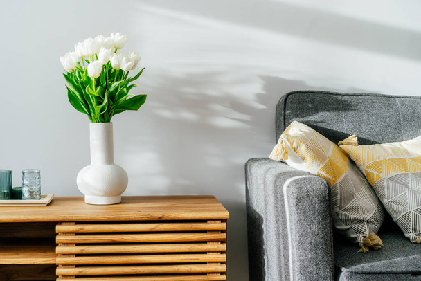 Scandinavian home interior with spring bouquet of white tulip flowers in glass vase, tray with candles standing on a wooden cabinet. Minimalist design with gray sofa and white wall. Springtime. - Фото, зображення