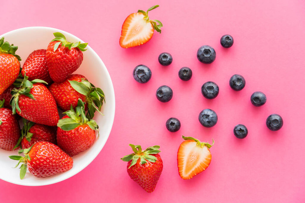 Top view of organic strawberries in bowl near blueberries on pink background  - Zdjęcie, obraz
