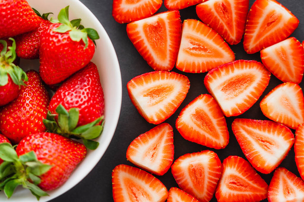 Flat lay with cut and whole strawberries in bowl on black background  - Foto, Imagen