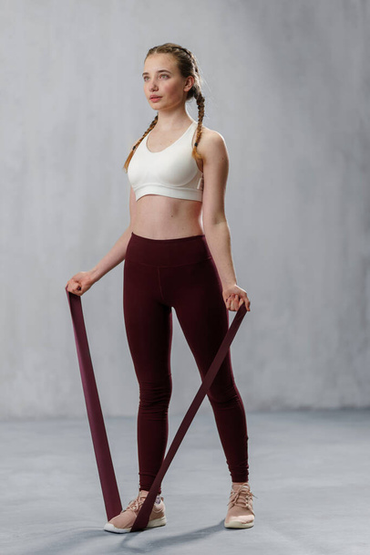 A sports woman in fashion sportswear exercising with elastic band stretch over greay background. - Фото, зображення