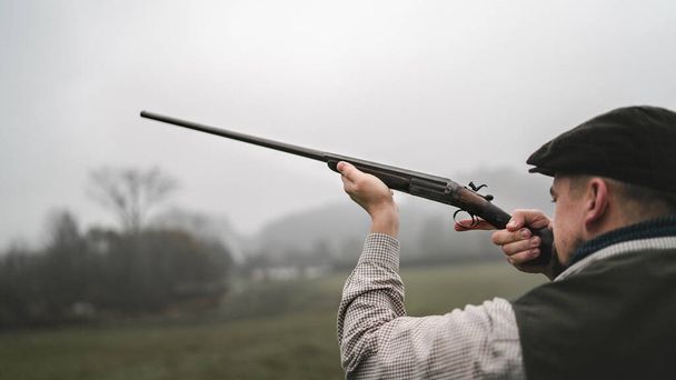 Hunter man in traditional shooting clothes on field aiming with shotgun. - Photo, Image