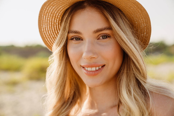 Young blonde woman wearing straw hat smiling and looking at camera on beach - Foto, immagini