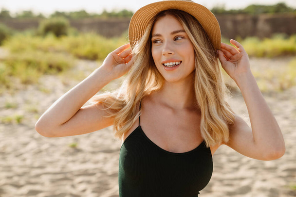 Mladá blondýna žena nosí slamák s úsměvem a dívá se stranou na pláži - Fotografie, Obrázek