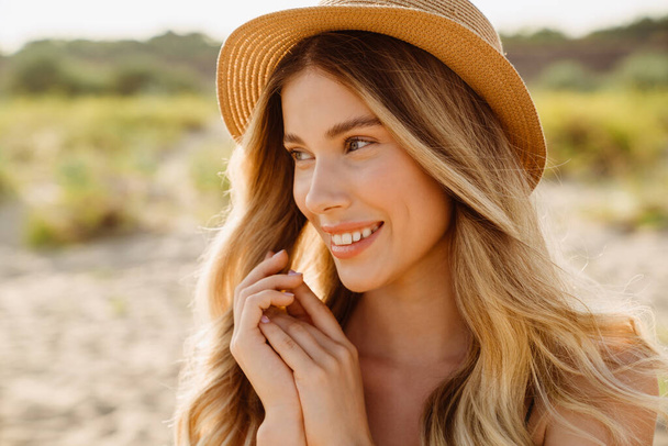 Νεαρή ξανθιά γυναίκα φοράει ψάθινο καπέλο χαμογελώντας και ατενίζοντας την παραλία - Φωτογραφία, εικόνα