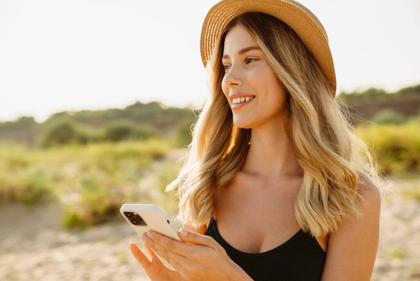 Young blonde woman wearing swimsuit smiling and using cellphone on beach - Photo, image