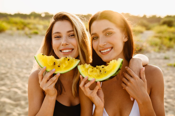 Young two women eating watermelon and hugging during picnic on beach - Photo, Image