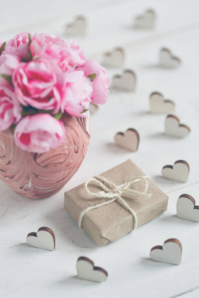 Piękna kompozycja z prezentem zawiniętym w papier kraft i delikatne róże w różowym wazonie na białym drewnianym tle. Wystrój wokół kompozycji z małymi drewnianymi sercami. - Zdjęcie, obraz
