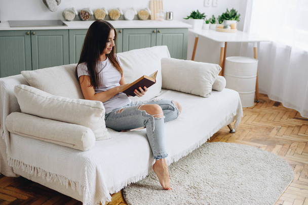 bruneta v roztrhaných džínách sedí v domácím interiéru na pohovce s vyplivnutými kostkami, uvolňuje se, odpočívá a pozorně čte román. Interiér s parketovou podlahou - Fotografie, Obrázek