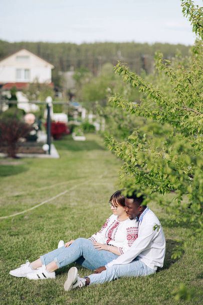 Fajok közötti pár ül a füvön a tavaszi kertben öltözött ukrán hagyományos etnikai hímzett ingek. A szerelmi kapcsolatok fogalma és a különböző emberi fajok közötti egység. - Fotó, kép