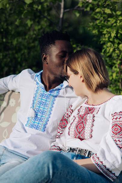 異人種間のカップルは、ウクライナの伝統的な民族刺繍のシャツを着た庭のベンチに座っています。異なる人種間の愛の関係と団結の概念. - 写真・画像