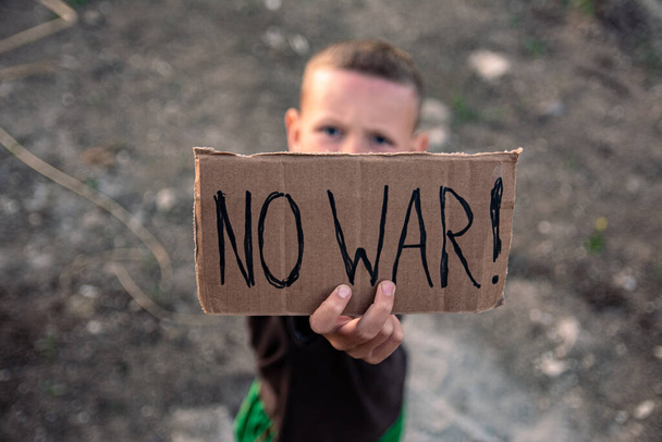 Un niño con la cara sucia y los ojos tristes sostiene un cartel de cartón con la inscripción NO GUERRA en inglés en sus manos. Un niño de apariencia europea se opone a la acción militar. - Foto, imagen