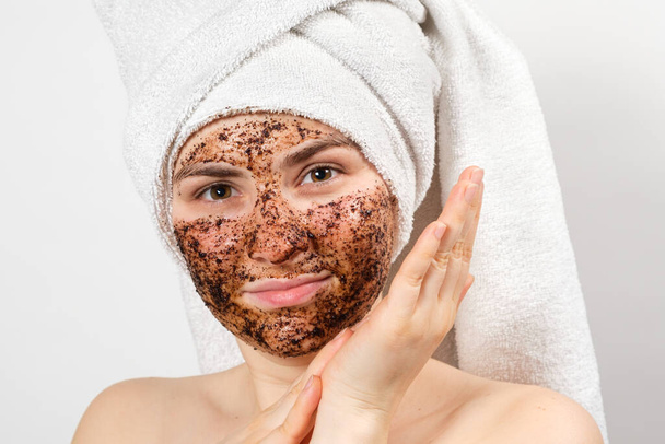 Piękna młoda kobieta z ręcznikiem kąpielowym na głowie i maską do kawy na twarzy dba o jej skórę.. - Zdjęcie, obraz
