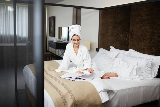 Χαμογελώντας νεαρή γυναίκα σε λευκό μπουρνούζια με πετσέτα στο κεφάλι hier, διαβάστε περιοδικά, στηρίζεται στο κρεβάτι. Οριζόντια προβολή. - Φωτογραφία, εικόνα