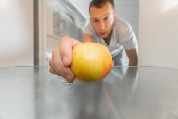 Man takes out a single apple from an empty refrigerator. Concept of delivery service, hunger, dients . Photo from inside the refrigerator - Photo, Image