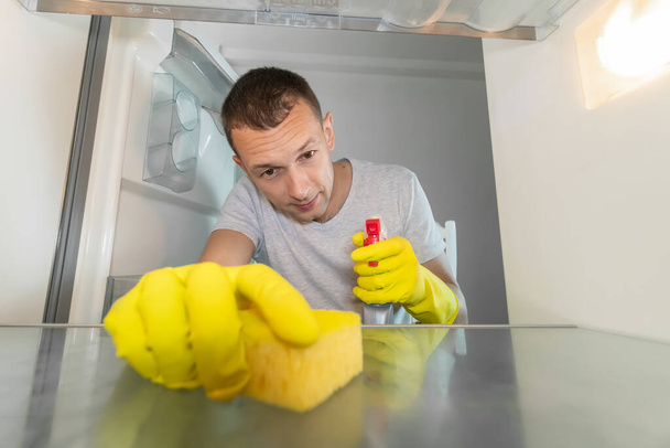 Lastik eldivenli bir adam buzdolabını sünger ve temizlik malzemesiyle temizliyor. Buzdolabının içinden fotoğraf. - Fotoğraf, Görsel