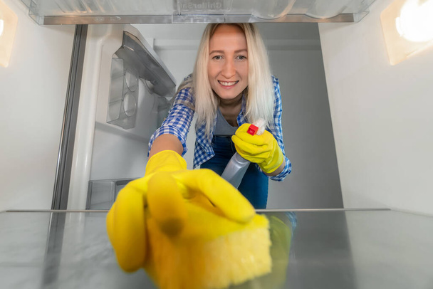 Hymyilevä nuori nainen keltaisissa kumikäsineissä pesee jääkaapin sisällä. Kuva jääkaapin sisältä. Siivouksen käsite, kotityöt - Valokuva, kuva