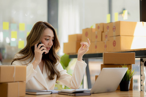 Asian woman is talking on the phone with a customer to confirm an order, she owns an online store, she packs and ships through a private transport company. Online selling and online shopping concepts. - Foto, imagen