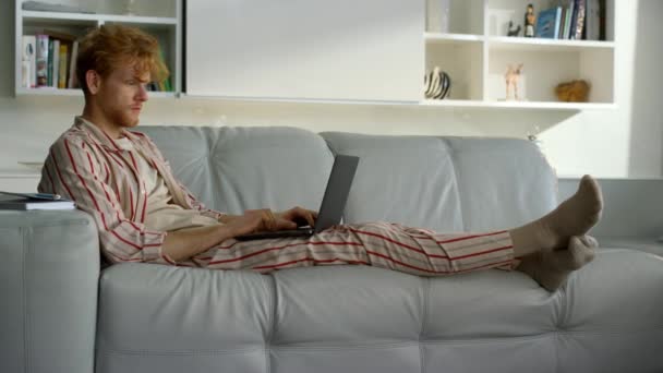 Человек, работающий на дому в пижаме. Сосредоточенный рыжий студент, использующий ноутбук, отдыхающий на диване. Серьезный фрилансер просматривает веб-смотреть онлайн вебинар на удаленном рабочем месте. Концепция карантинного образа жизни - Кадры, видео