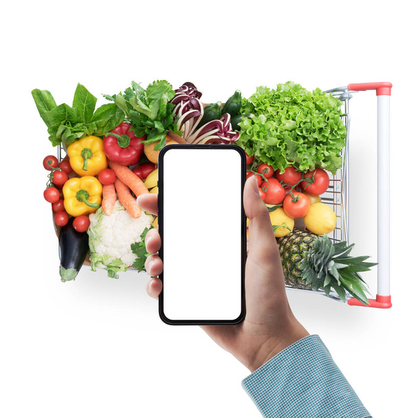 Customer holding a smartphone with blank screen and shopping cart full of fresh vegetables and fruits Isolated on white background - Photo, Image