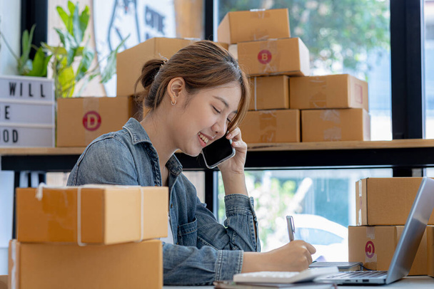 Asian woman is talking on the phone with a customer to confirm an order, she owns an online store, she packs and ships through a private transport company. Online selling and online shopping concepts. - Fotoğraf, Görsel