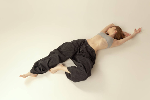 One slim girl lying on floor in top and giant black jeans, posing isolated over grey studio background. Weight-loss. Concept of beauty, health, body and skin care, spa, cosmetology, fitness, ad - Фото, изображение