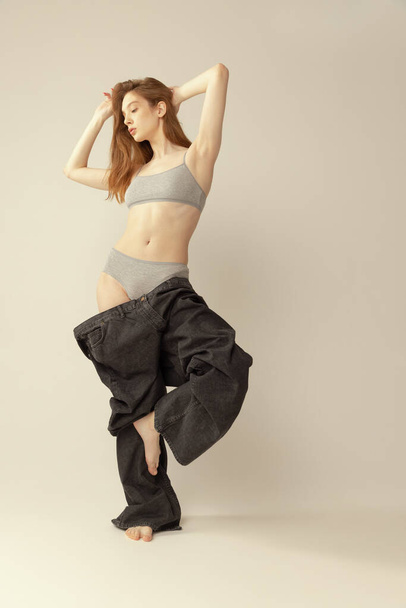 Portrait of young slim girl in top and giant black jeans posing isolated over grey studio background. Weight-loss. Concept of beauty, health, body and skin care, spa, cosmetology, fitness, ad - 写真・画像