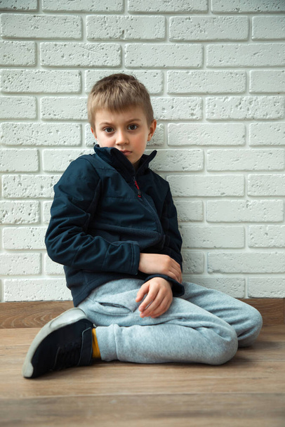 Ένα σκεπτικό αγόρι 6-7 ετών κάθεται στο πάτωμα με ένα μυστηριώδες βλέμμα. Πορτραίτο ενός αγοριού, μπροστινή όψη - Φωτογραφία, εικόνα