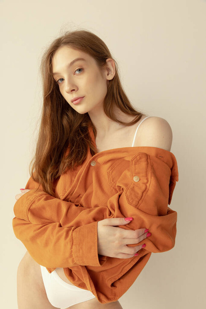 Portrait of beautiful red-haired young girl posing in orange shirt and underwear isolated over grey studio background. Concept of beauty, health, body and skin care, spa, cosmetology, fitness, ad - 写真・画像