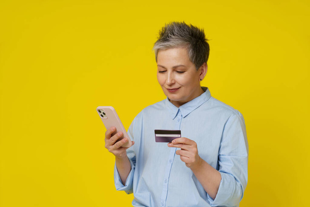 Mature grey haired woman hold smartphone and debit or credit card in hand making online payment or shopping. Pretty woman in blue shirt buying online isolated on yellow background. Focus on the phone. - Photo, image