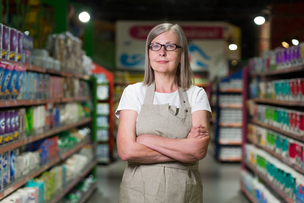Muotokuva vanhempi nainen lasit johtaja ruokakauppa, supermarket. Seisoo työvaatteissa, kädet ristissä, katsoo kameraan, hymyilee - Valokuva, kuva
