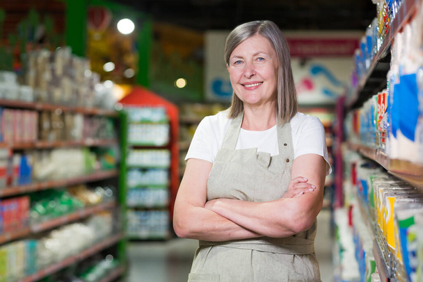 Портрет старшої жінки-консультантки, співробітника в продуктовому магазині, супермаркеті. Стоячи в робочому одязі, руки схрещені, дивлячись на камеру, посміхаючись
 - Фото, зображення