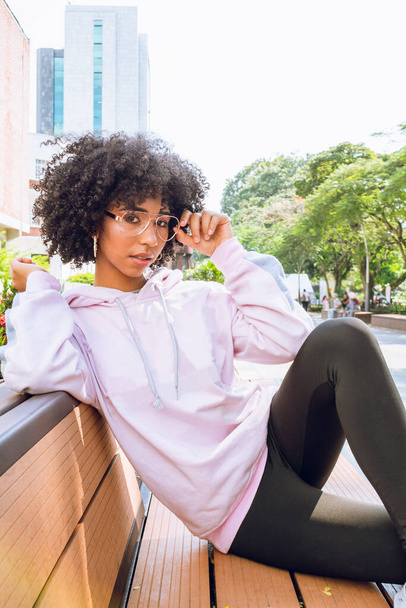 красива молода афроамериканка сидить на лавці модель позує насолоджуючись сонячним днем у громадському міському парку
 - Фото, зображення