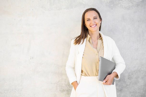Confident young business woman carrying laptop standing over gray wall, smiling proud female office employee in white smart casual suit looking at the camera - Φωτογραφία, εικόνα