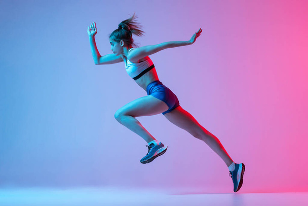 Studio shot of professional female athlete, runner training isolated on pink studio background with blue neon filter, light. Concept of action, motion, speed, healthy lifestyle. Copy space - 写真・画像