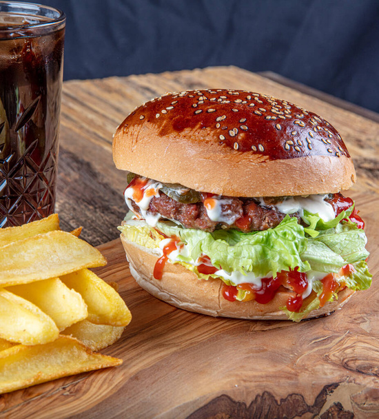 Sappige en smakelijke hamburger met tomaten, sla, komkommer, gesmolten kaas en uien met brioche broodje geserveerd op een houten bord met frietjes - Foto, afbeelding