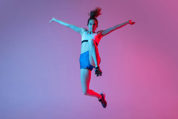 Moments. Young girl, female athlete training long jump technique isolated on pink studio background with blue neon filter, light. Concept of action, motion, speed, healthy lifestyle. Copy space - Zdjęcie, obraz