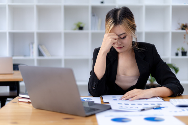 Een zakenvrouw heeft hoofdpijn na een lange tijd voor haar laptop te hebben gewerkt en als ze naar veel papierwerk kijkt dat stress veroorzaakt, rust ze en werkt ze hard. Kantoorsyndroom concept. - Foto, afbeelding