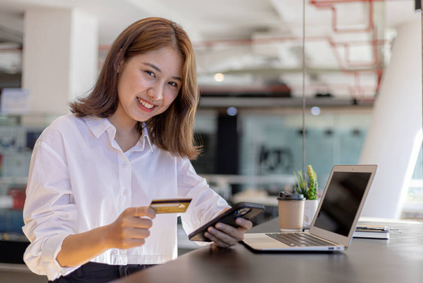 A woman sitting at the counter holding a cell phone and credit card, she is shopping online on a smartphone, she pays for goods and services by credit card. Concept of using credit card for payment. - 写真・画像