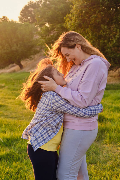 Πλευρική άποψη για χαρούμενη μητέρα και κόρη αγκαλιάζει ο ένας τον άλλον, παίζοντας στο πάρκο στο πράσινο γρασίδι την άνοιξη ηλιόλουστη μέρα, ευτυχισμένη οικογενειακή ζωή - Φωτογραφία, εικόνα