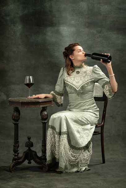 Tasting red wine. Conceptual portrait of young charming girl in image of medieval royal person or viscountess drinking wine isolated on dark background. Comparison of eras concept, fashion, art, ad. - Фото, изображение